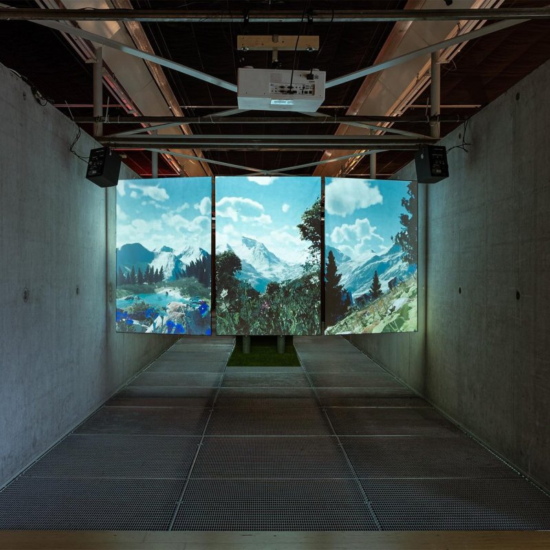 Installation view: a dark concrete hall with a three-panel projection of an alpine landscape at the far end