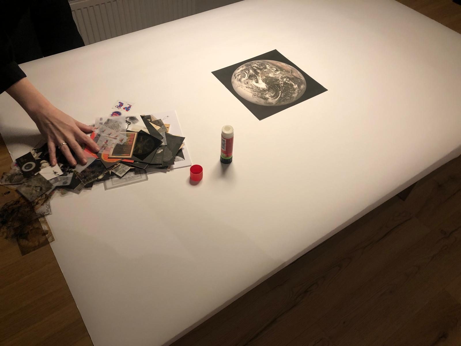 A hand arranging cut-outs on a white table next to a black-and-white print of the Earth and a glue stick