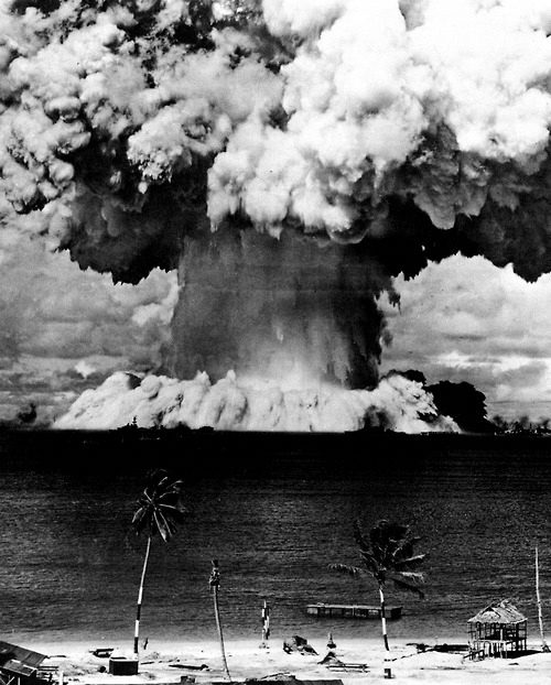 Black-and-white photograph of a nuclear test explosion rising above a beach with palm trees