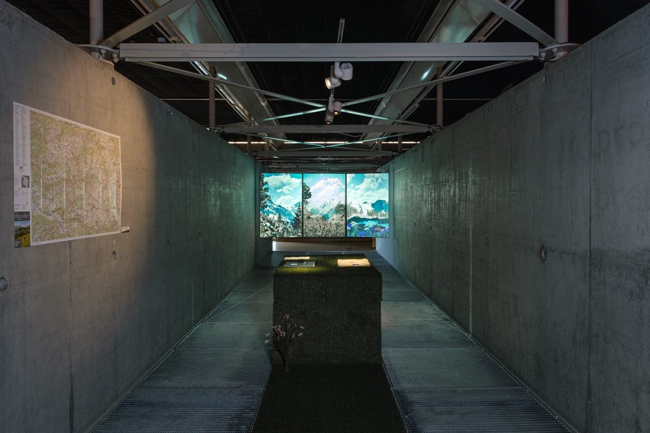 View down the long concrete hall towards the projected landscape, with a map pinned to the left wall